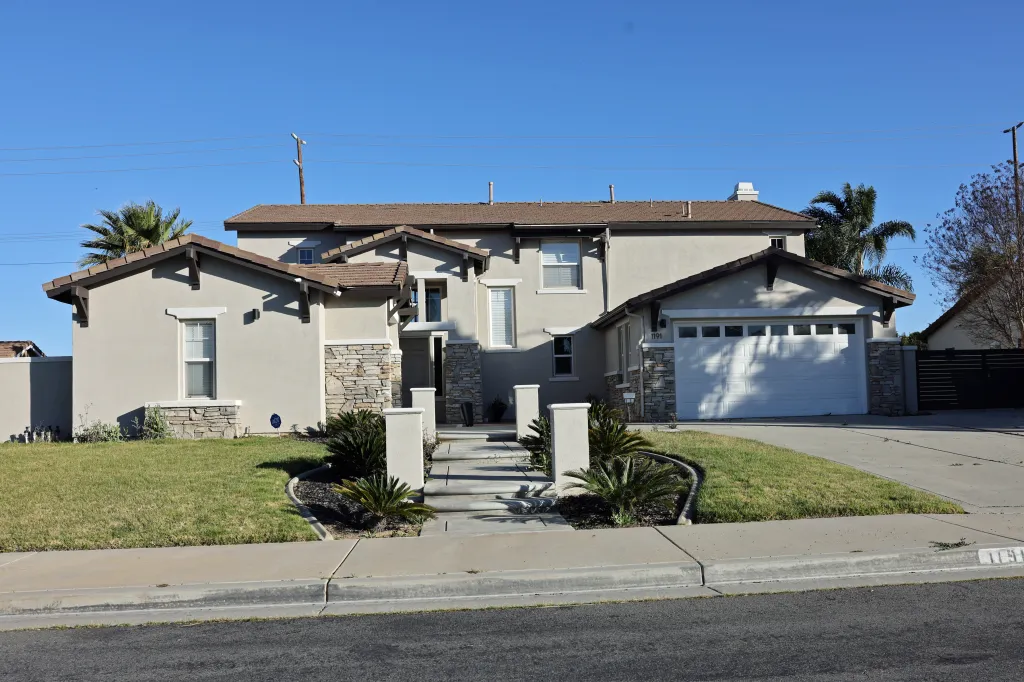 Two-story house at 1191 Pamplona Dr. in Riverside, CA, featuring a tan exterior with stone accents, a two-car garage, and visible security cameras.