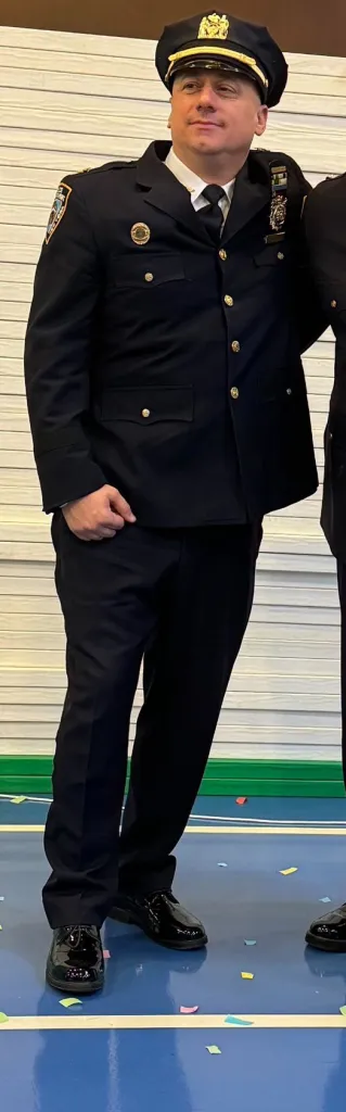 A man in a police uniform, with a black cap, dark blue jacket with gold buttons, and dark blue trousers, stands on a blue floor with colorful confetti.