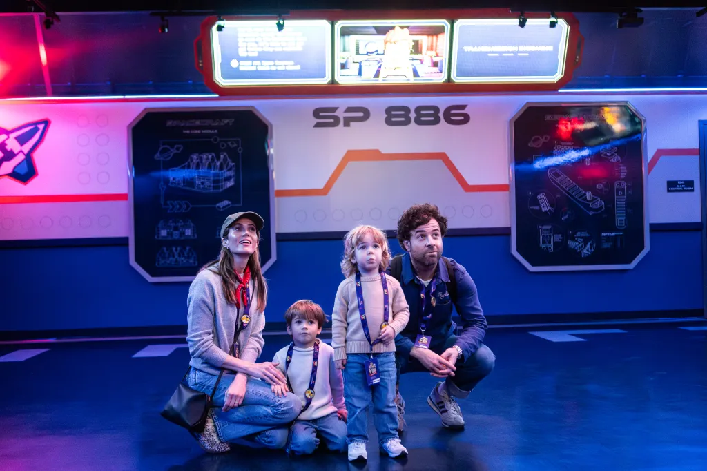 A family of four, including a mother, father, and two young children, kneel in a brightly lit, futuristic-looking room with blue and pink lighting, wearing VIP badges around their necks and looking up intently.