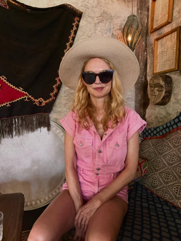 A woman in a pink romper, straw hat, and sunglasses sitting on a patterned cushion.