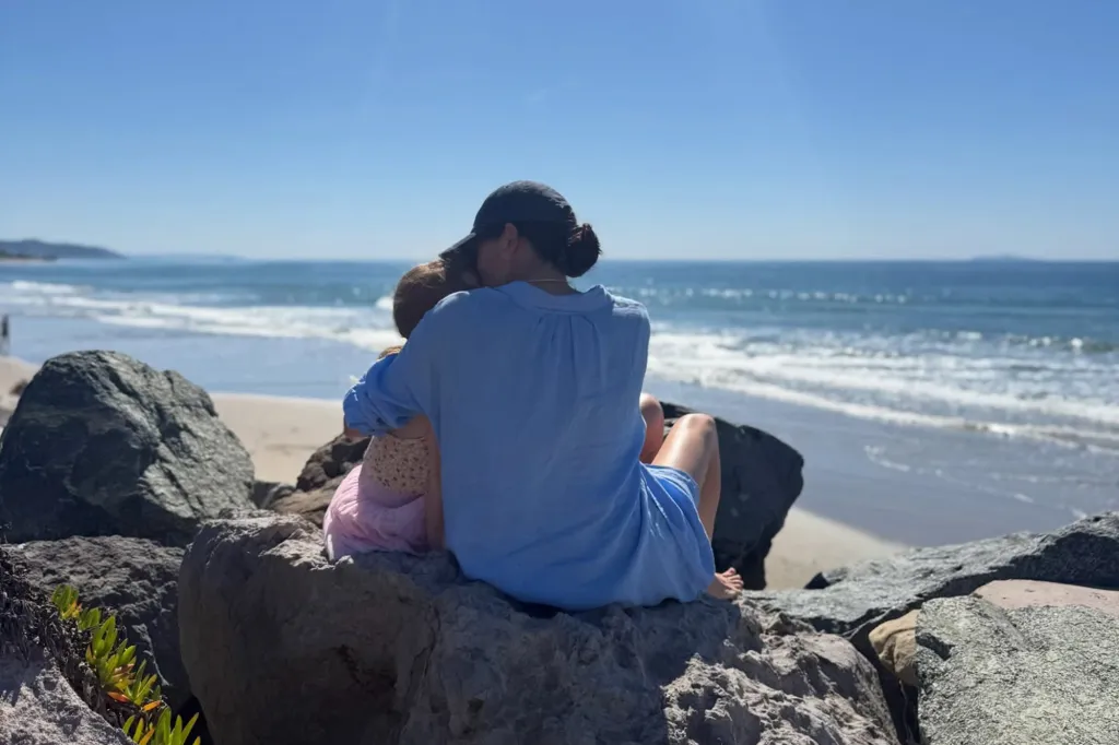 A person in a blue shirt with a child in a pink outfit sitting on rocks looking at the ocean.