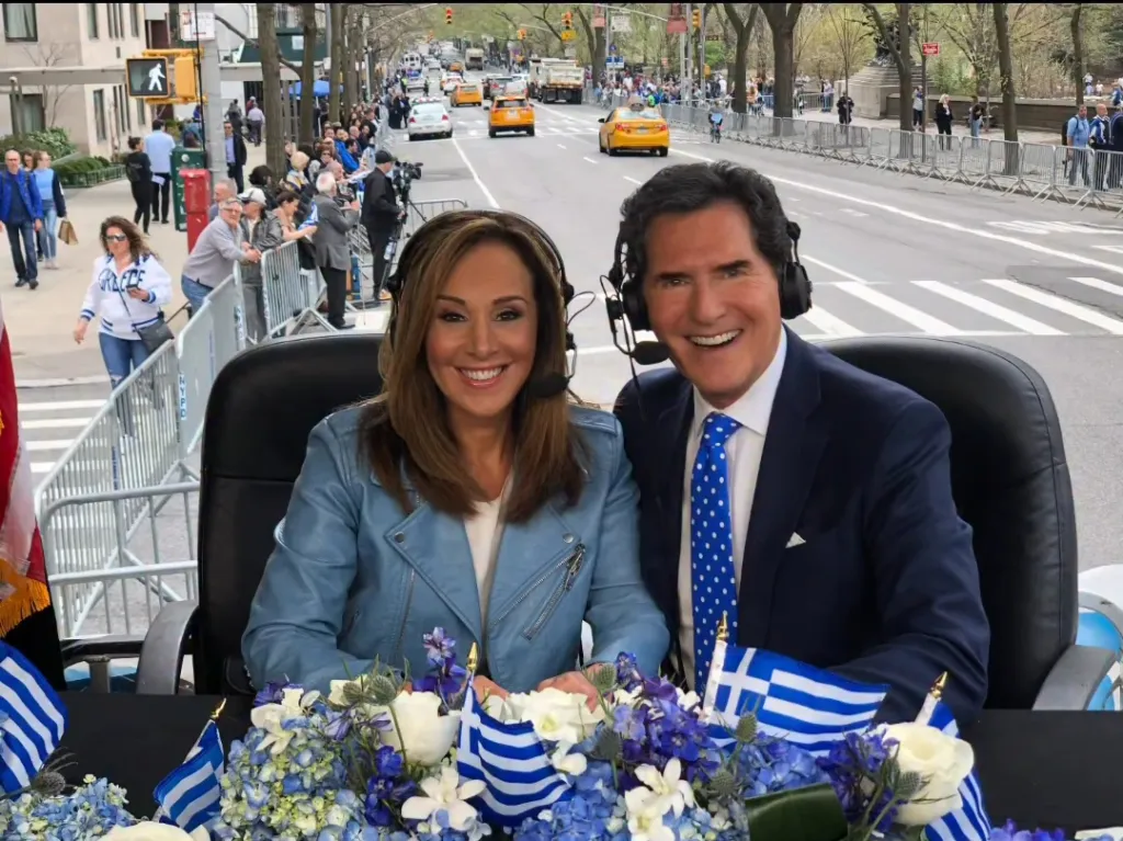 Two news anchors with headsets sit behind a table decorated with Greek flags and flowers, with a parade in the background.
