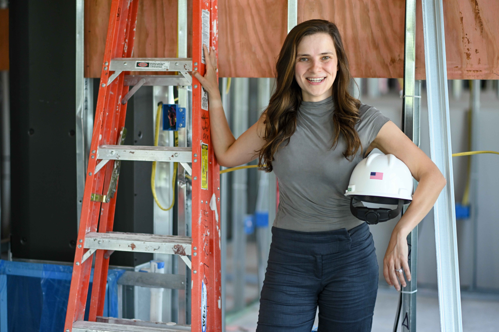 Smiling woman in work clothes holding a hardhat, with a construction ladder in the foreground.