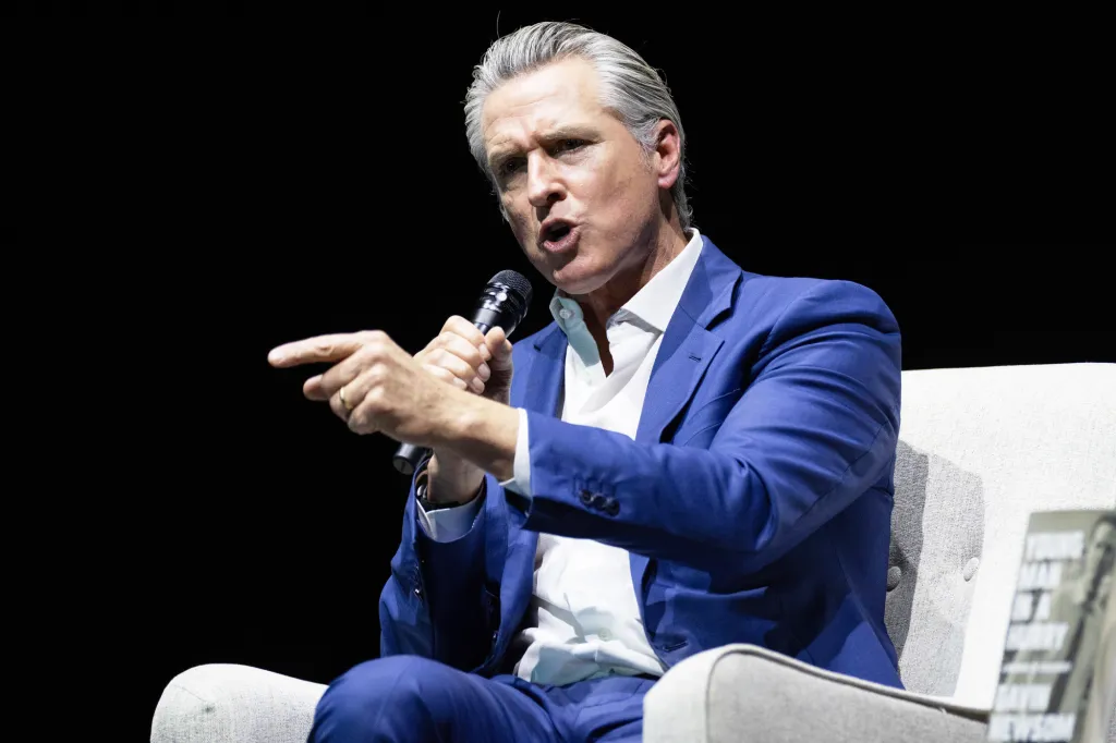 Gavin Newsom speaks into a microphone while sitting in a chair.