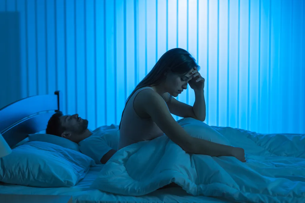 A woman sitting up in bed, looking distressed, while a man sleeps beside her.