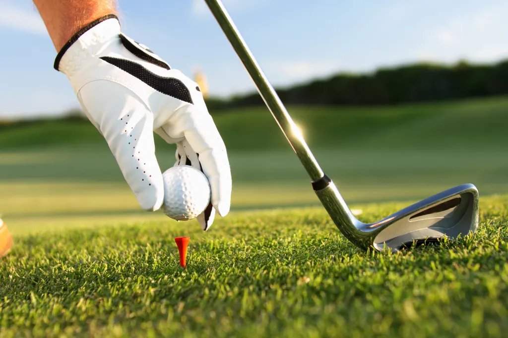 A golfer's gloved hand places a golf ball on a red tee, next to a golf club, on a green golf course.