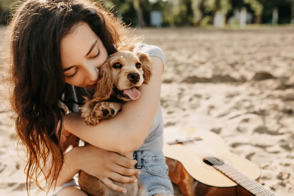 A young woman sitting on a beach hugging a Cocker Spaniel puppy.