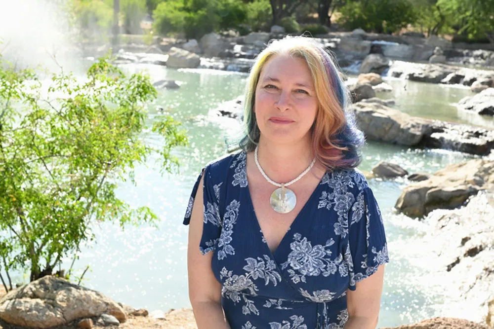 Woman with colorful hair and a patterned dress wearing a circular pendant stands by a body of water with rocks and trees.