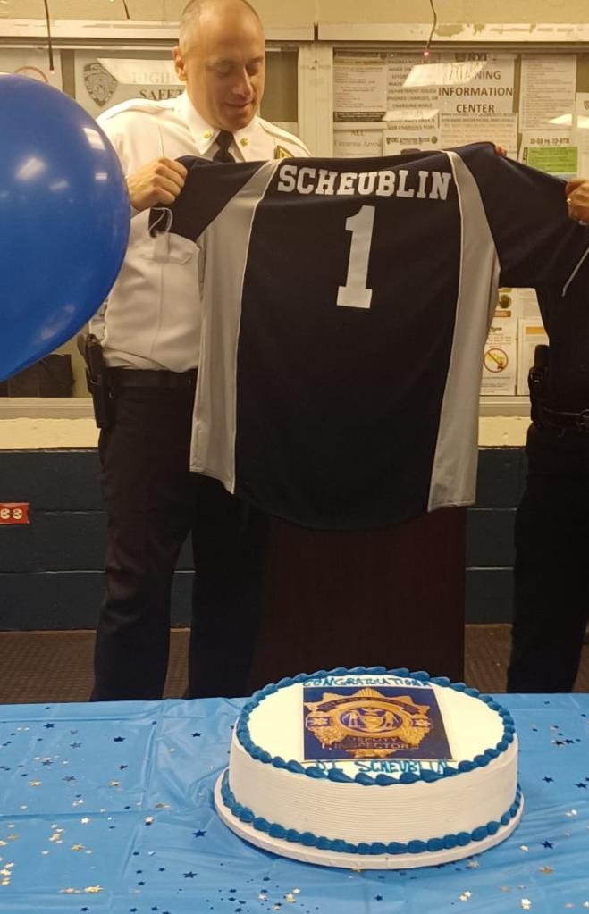 Police officer Jeremy Scheublin holding up a jersey with his last name and number 1, with a celebratory cake in the foreground.