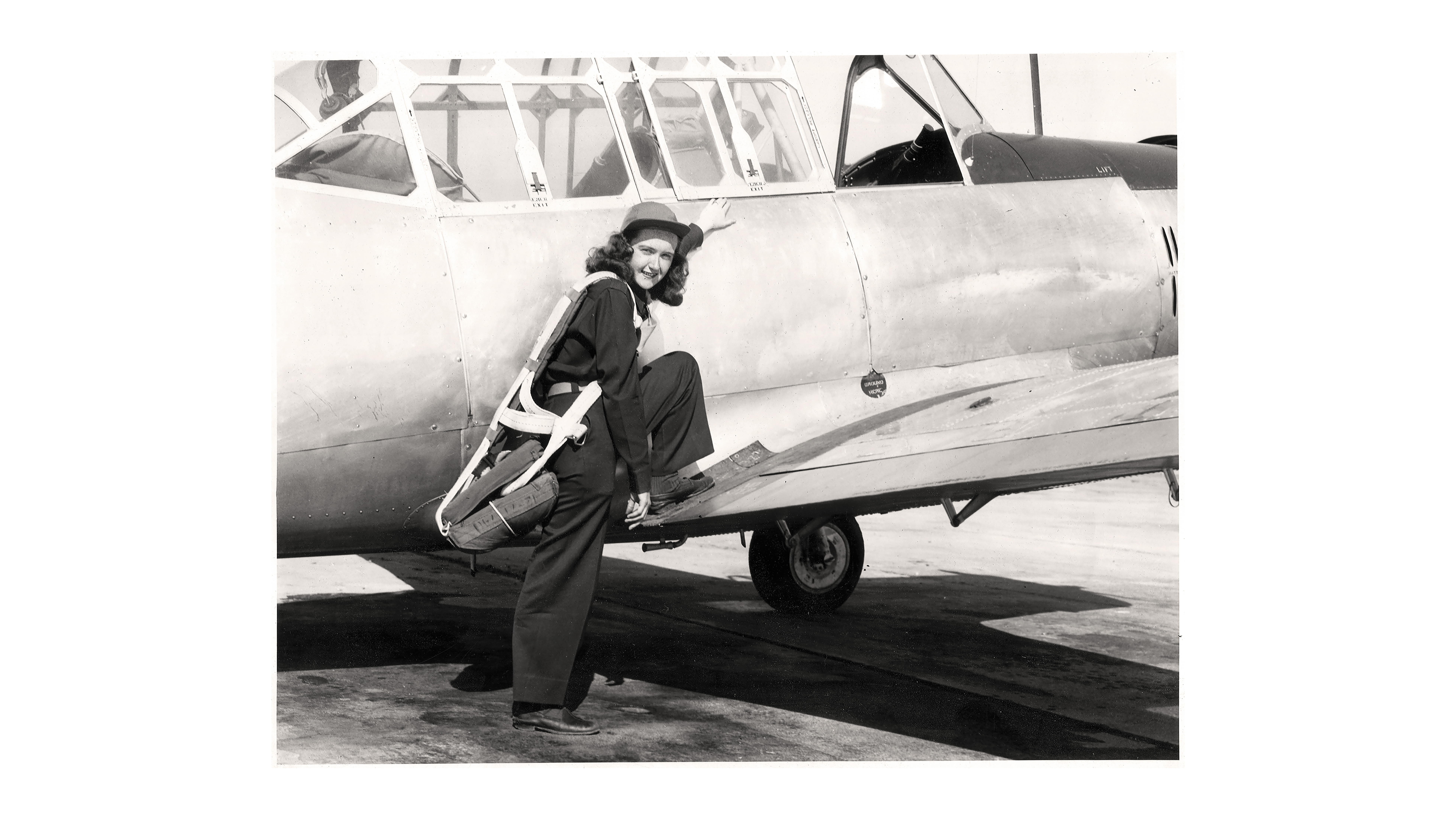 black-and-white archival photo of woman in flight gear and cap, smiling at camera, standing with one foot on ground and one foot on the wing of the plane she piloted, about to climb into cockpit