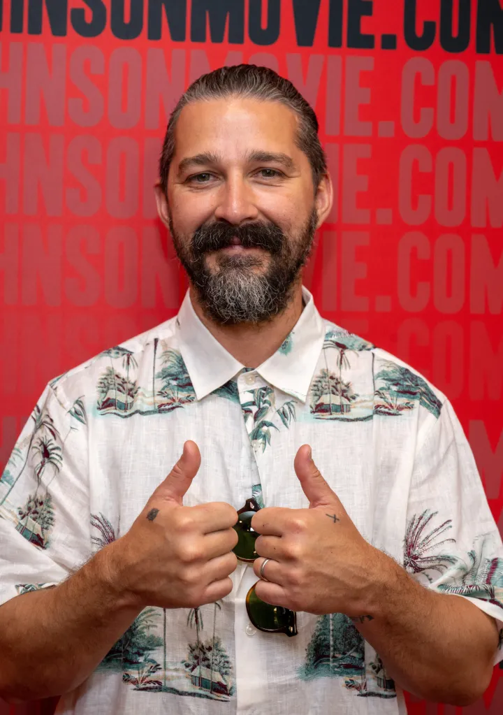 Shia LaBeouf with a beard giving two thumbs up while wearing a Hawaiian shirt.