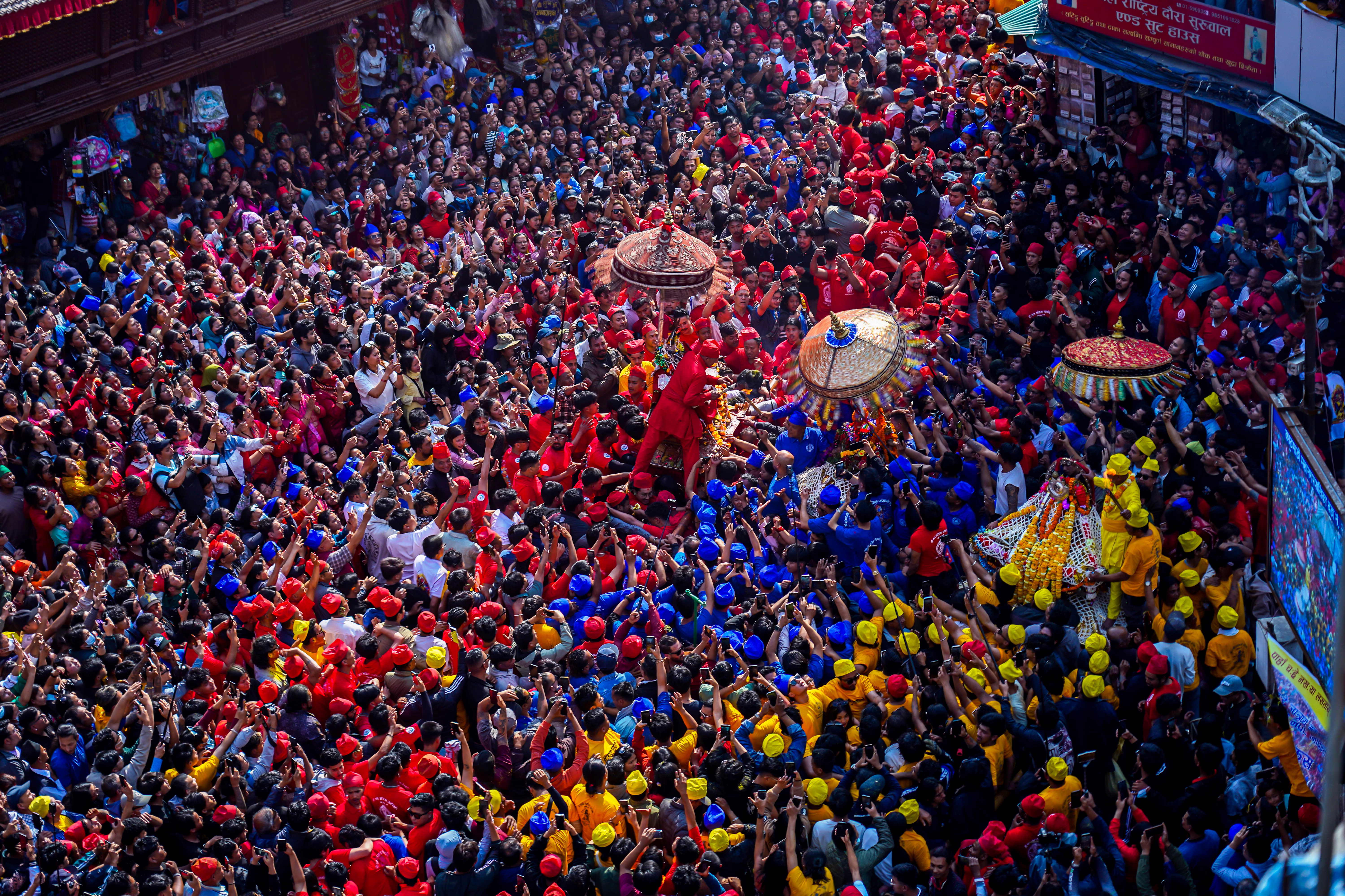 People wearing colorful costumes try to pull chariots through a densely-packed crowd.