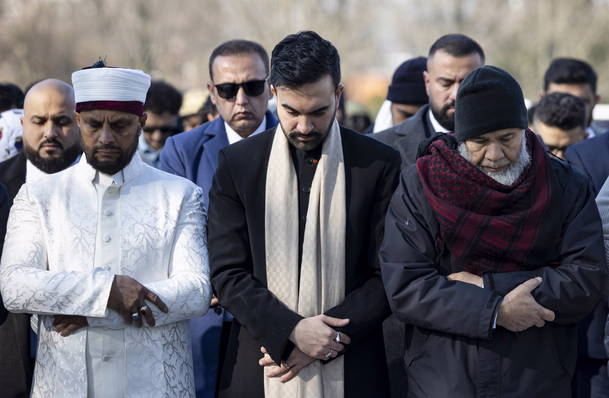 A group of men stand shoulder to shoulder, praying in a park, with New York City Mayor Zohran Mamdani standing at center.