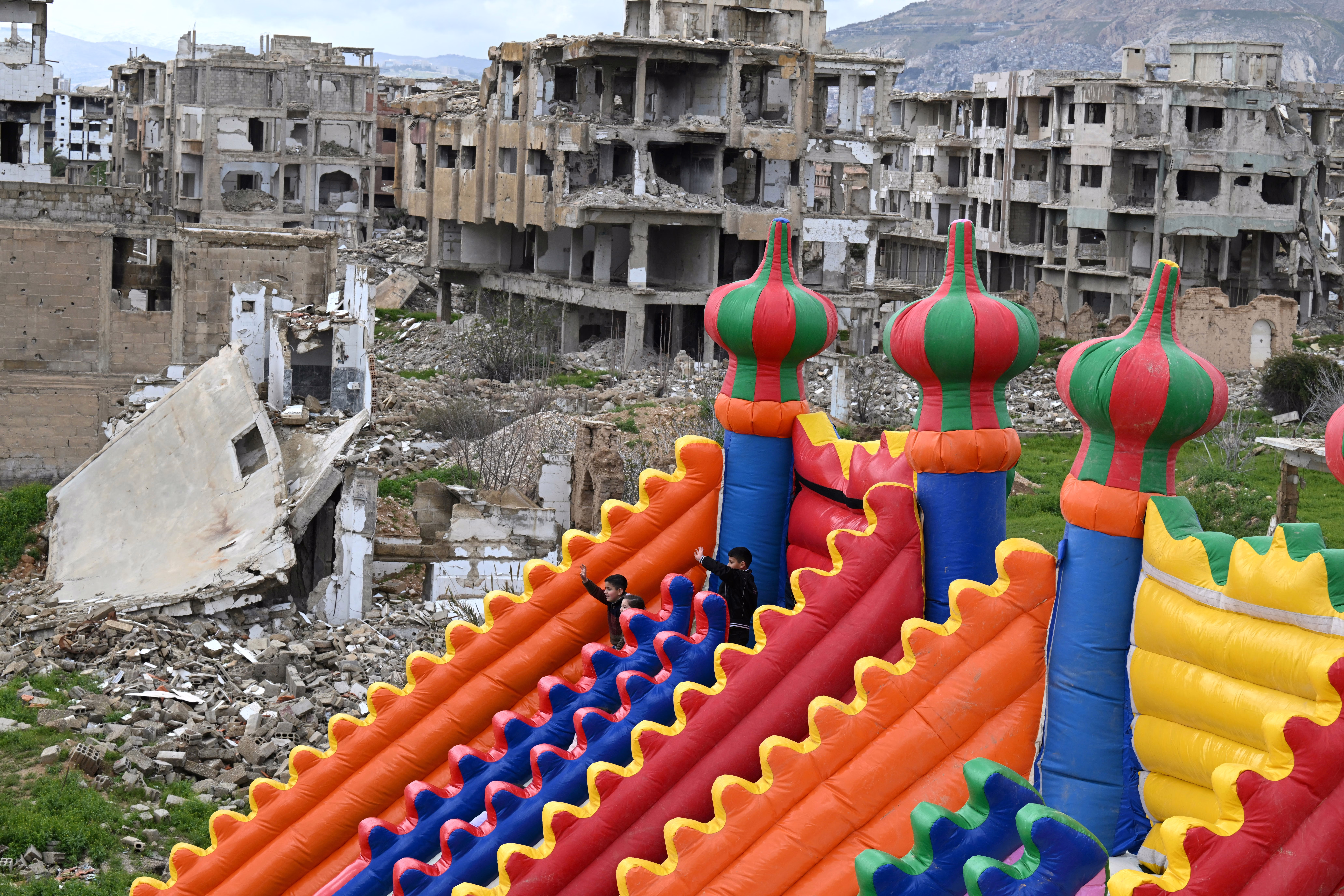Children enjoy rides at an amusement park set up on land surrounded by war-damaged buildings.