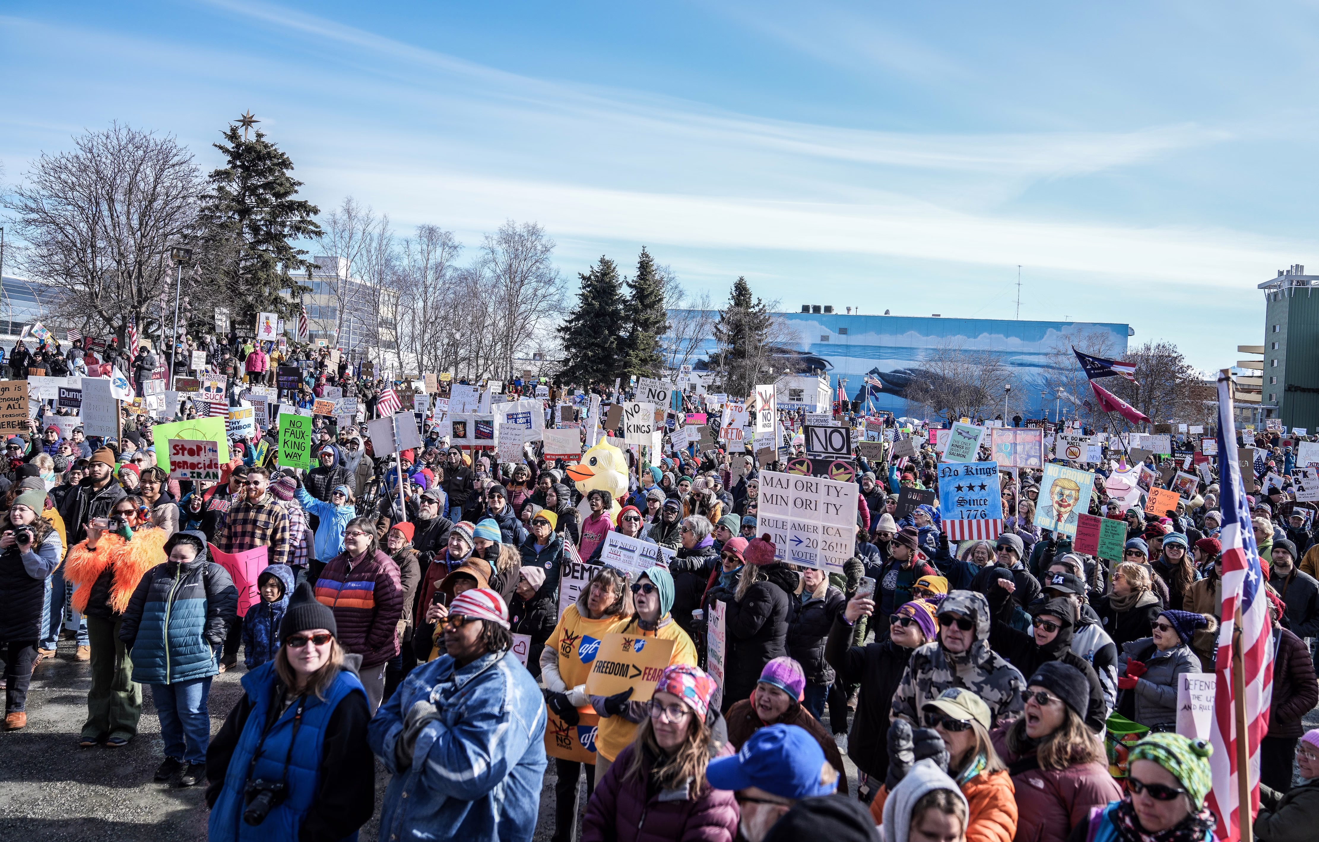 A group of protesters wearing warm clothing stand together in a city square holding signs