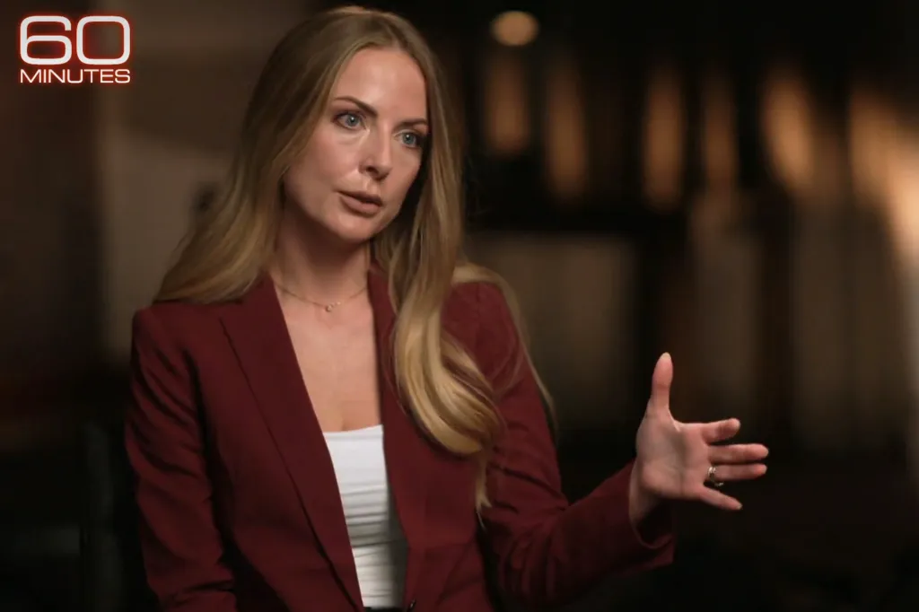 A blonde woman in a red blazer speaks to the camera, gesturing with her right hand.