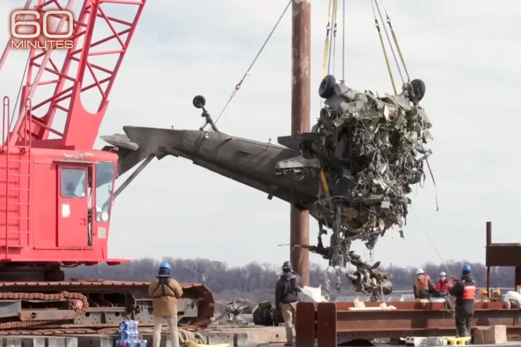 A red crane lifts the mangled fuselage of a helicopter from a body of water, with several workers watching from a barge.