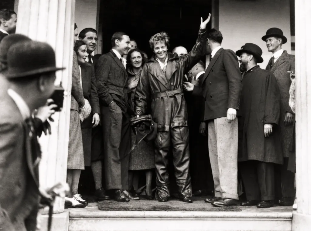 Amelia Earhart, dressed in a flight suit, smiling while surrounded by people in London after her solo trans-Atlantic flight.