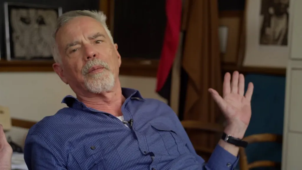 Robert Trivers, an older man with gray hair and a beard, wearing a blue checkered shirt and a lavalier microphone, gesturing with his hands.