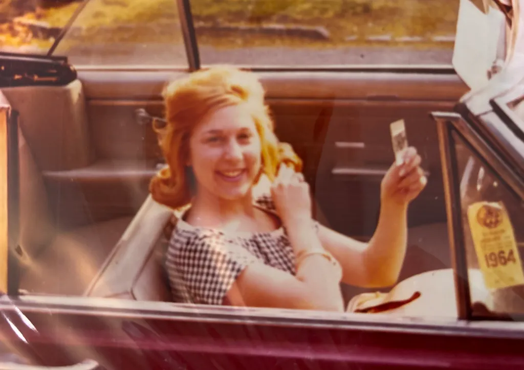 Barbara Waldman smiling while holding a small photo, seated in a car with a 1964 sticker on the windshield.