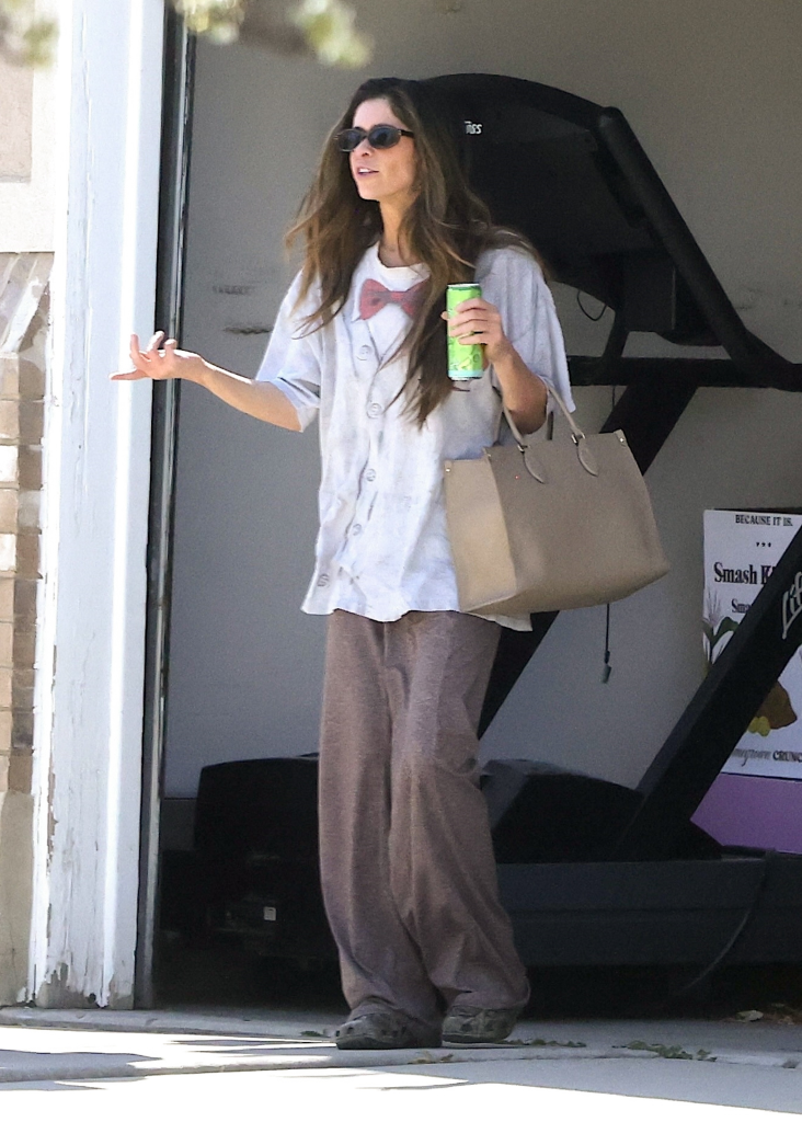 Taylor Frankie Paul in casual clothes and sunglasses, holding a drink and a large tote bag, standing outside with a treadmill in the background.
