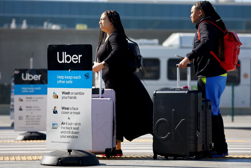 Travelers with luggage wait by an Uber sign with safety guidelines at Los Angeles International Airport.