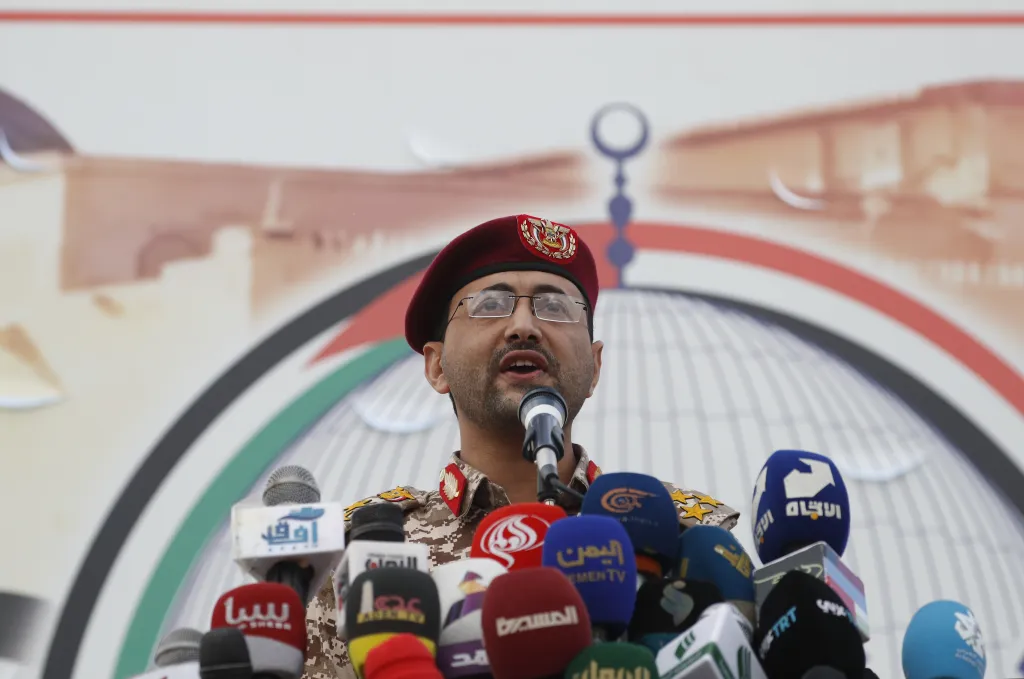 A man in a military uniform and a maroon beret speaks at a press conference, surrounded by microphones from various news outlets.