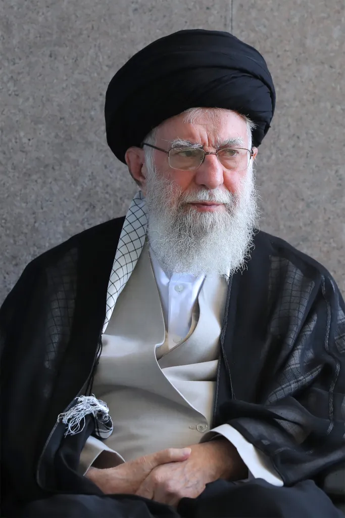 Ayatollah Seyyed Ali Khamenei, Leader of the Islamic Revolution, with a white beard and black turban, wearing a black robe over a beige vest and white shirt.