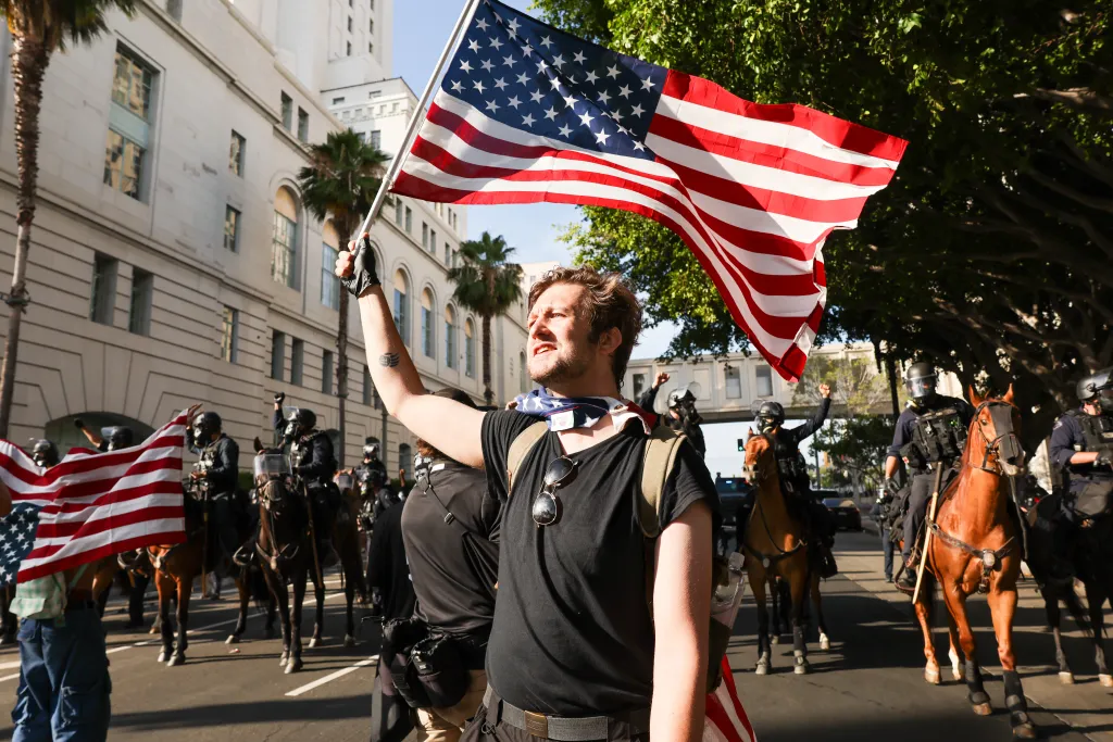 A man holding an American flag at an anti-Trump protest in Los Angeles, with mounted police in the background.