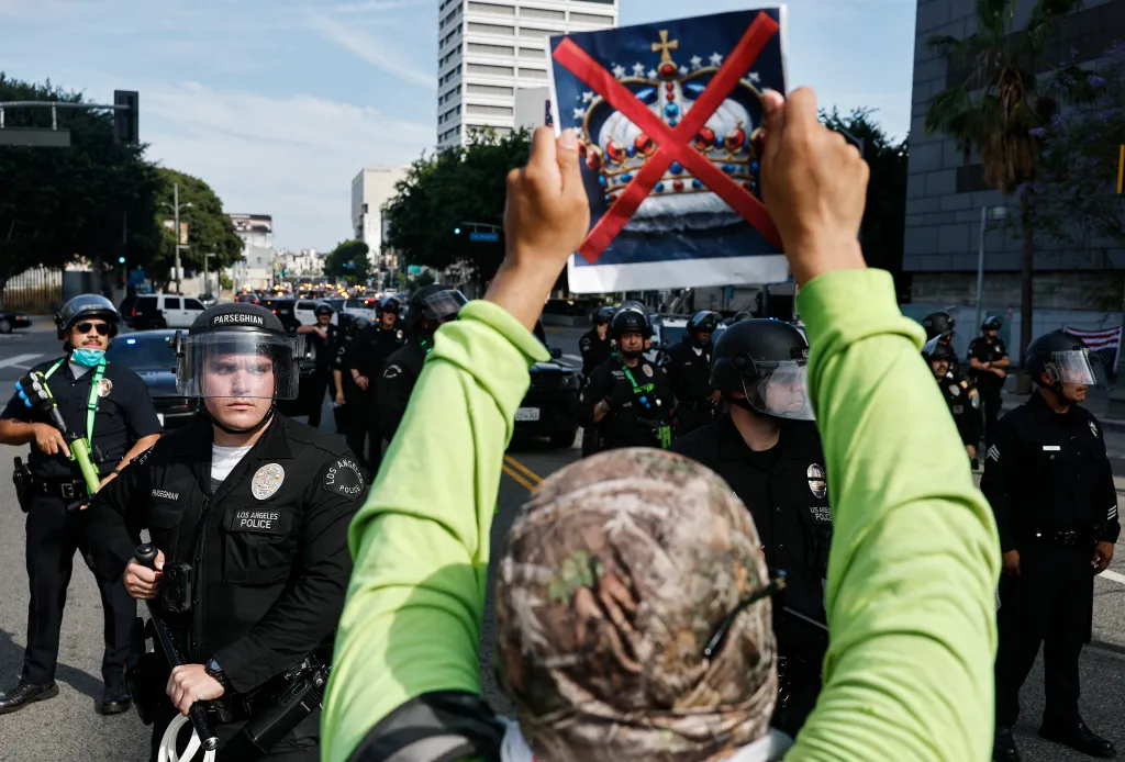 A protester holds a sign with a crown and a red X over it, facing Los Angeles Police Department officers.