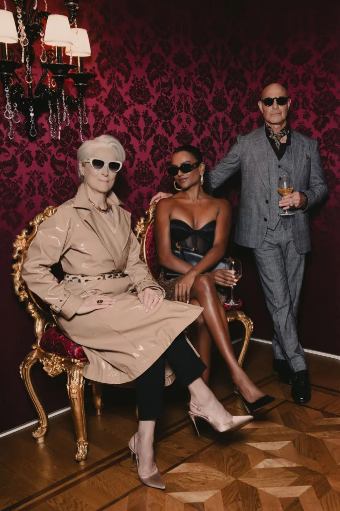 Meryl Streep in a beige vinyl trench coat and sunglasses, Simone Ashley in a black corset top and crystal mesh skirt and sunglasses, and Stanley Tucci in a grey wool suit and sunglasses at Milan Fashion Week.