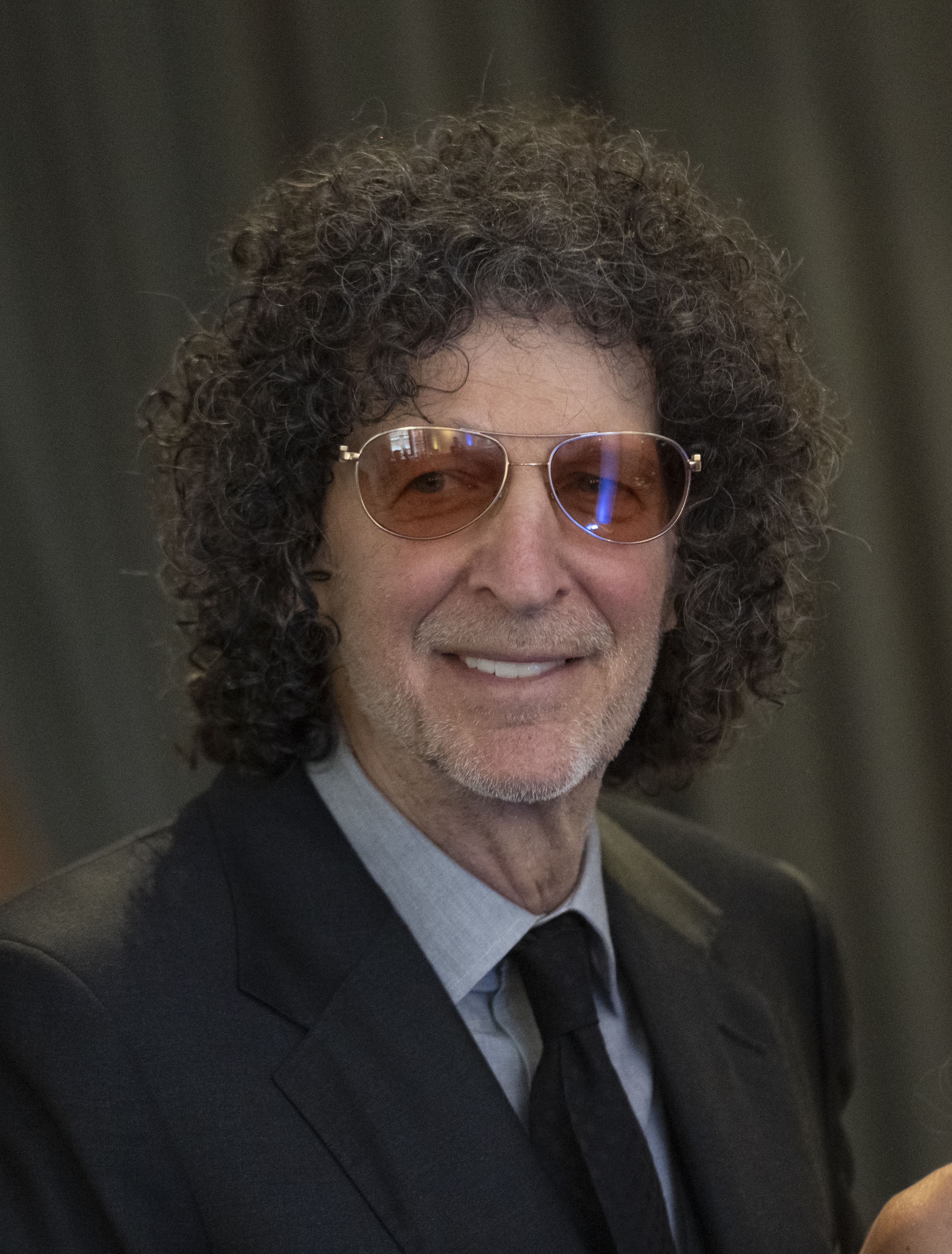 Howard Stern smiling in a suit and sunglasses.