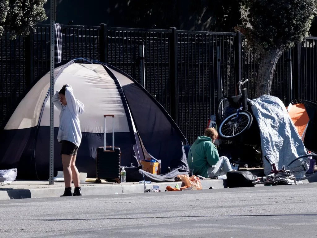 Homeless encampment in Los Angeles, featuring multiple tents, bicycles, and personal belongings lining a sidewalk next to a fence.
