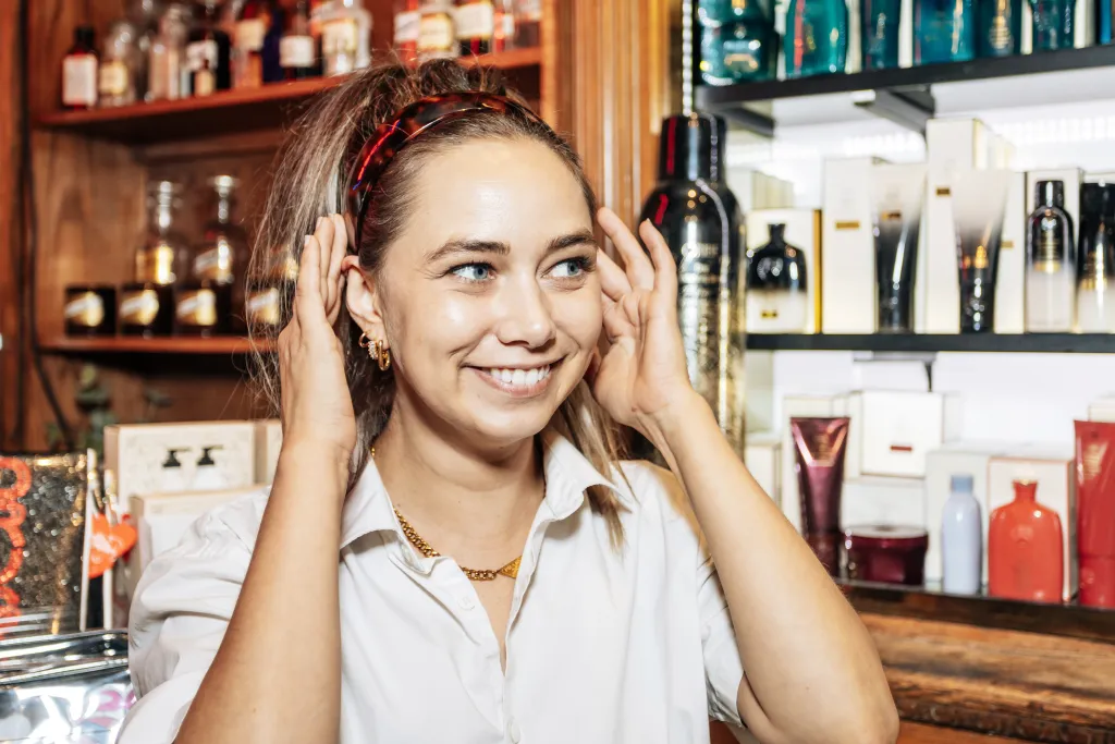 A smiling woman with light hair and blue eyes, wearing a white collared shirt and a headband, stands in front of shelves of cosmetic products.