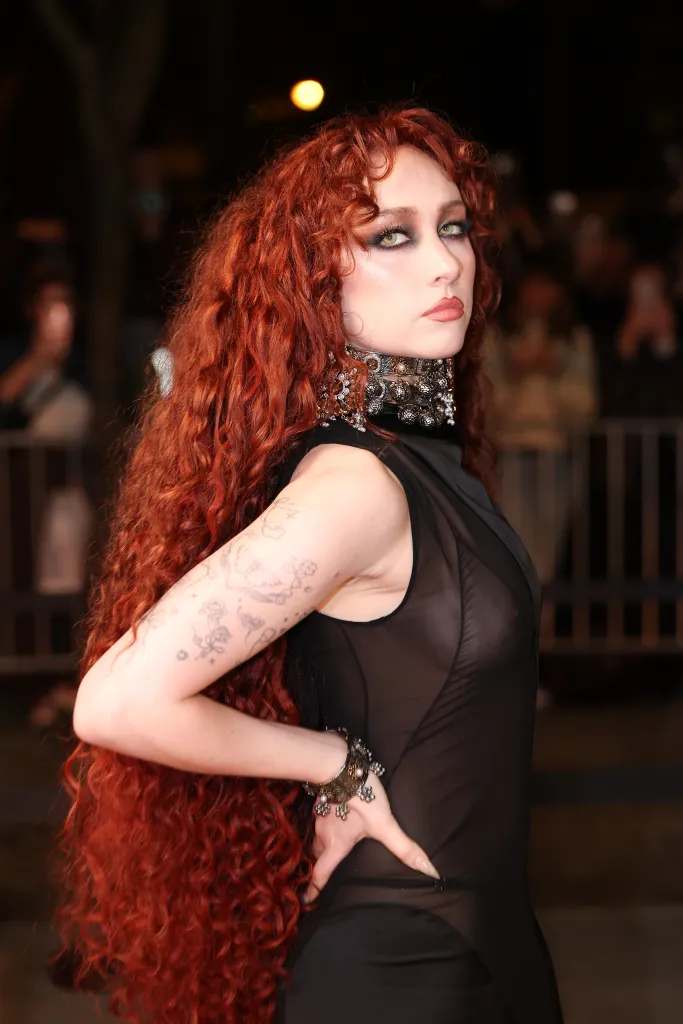 Chappell Roan with long red curly hair and dark eye makeup, posing in a sheer black outfit and ornate choker.