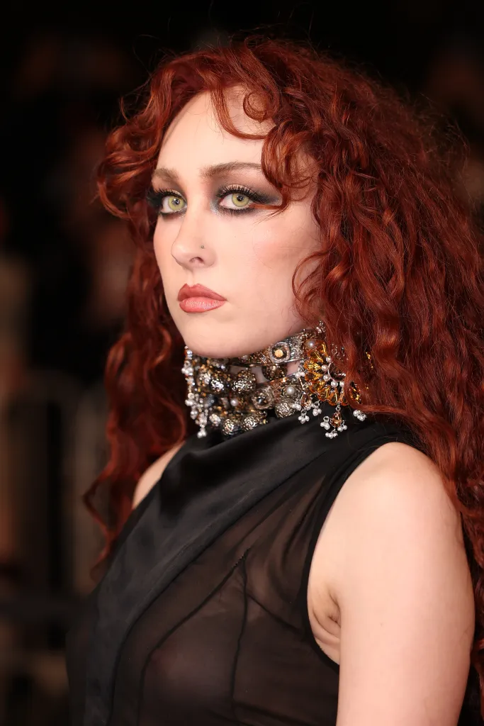 Chappell Roan at Paris Fashion Week in a black sheer top, elaborate collar necklace, and curly red hair.