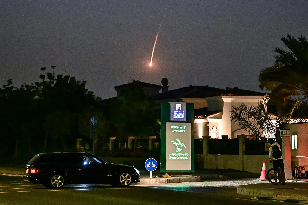 A bright projectile streaks across a dark night sky over Dubai, with a car, a sign for 