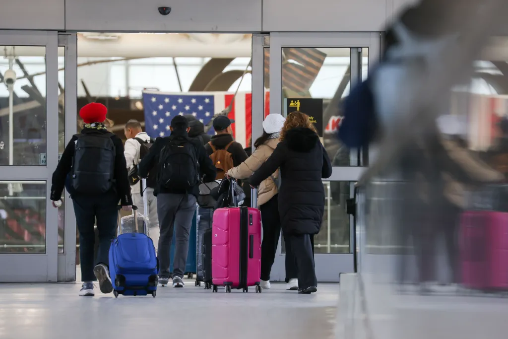Travelers with luggage enter John F. Kennedy International Airport (JFK) Terminal 1, with an American flag visible in the background.