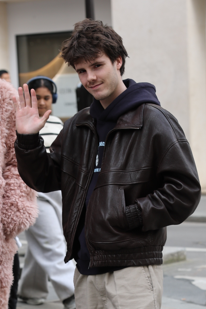 Cruz Beckham waves at the camera while wearing a brown leather jacket over a navy hoodie.