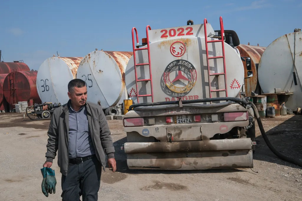 A worker at an oil stock market in Erbil, Iraq, holds work gloves with large, rusted oil tanks and a tanker truck in the background.