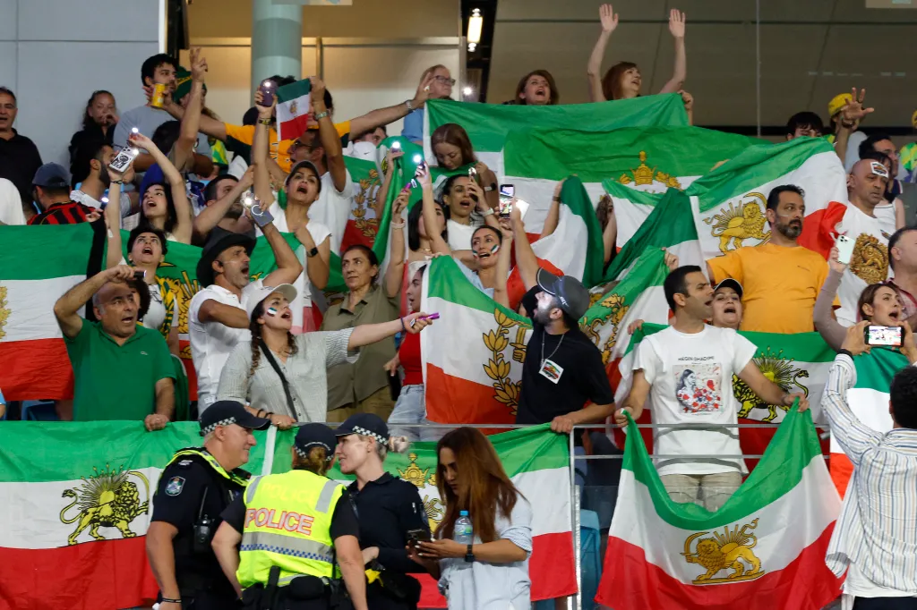 Iranian fans waving Pahlaviera flags and holding up cell phones with their flashlights on, with Australian police standing guard in the foreground.