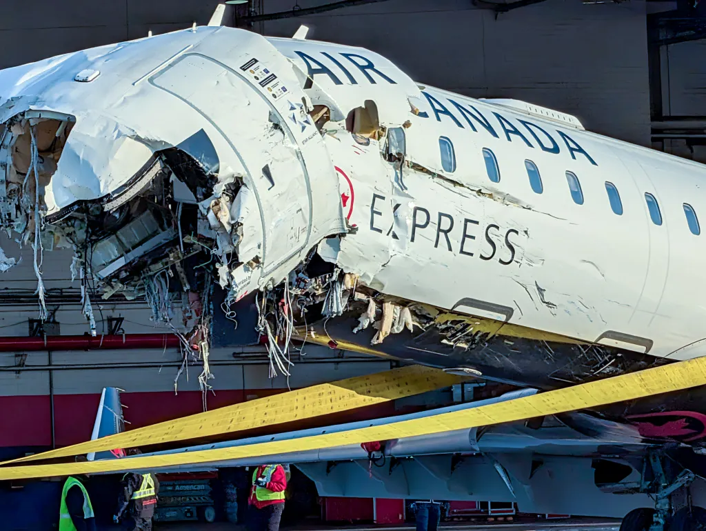 An Air Canada Express plane with significant damage to its front fuselage, including large torn sections, sits inside a hanger with yellow straps around its body.