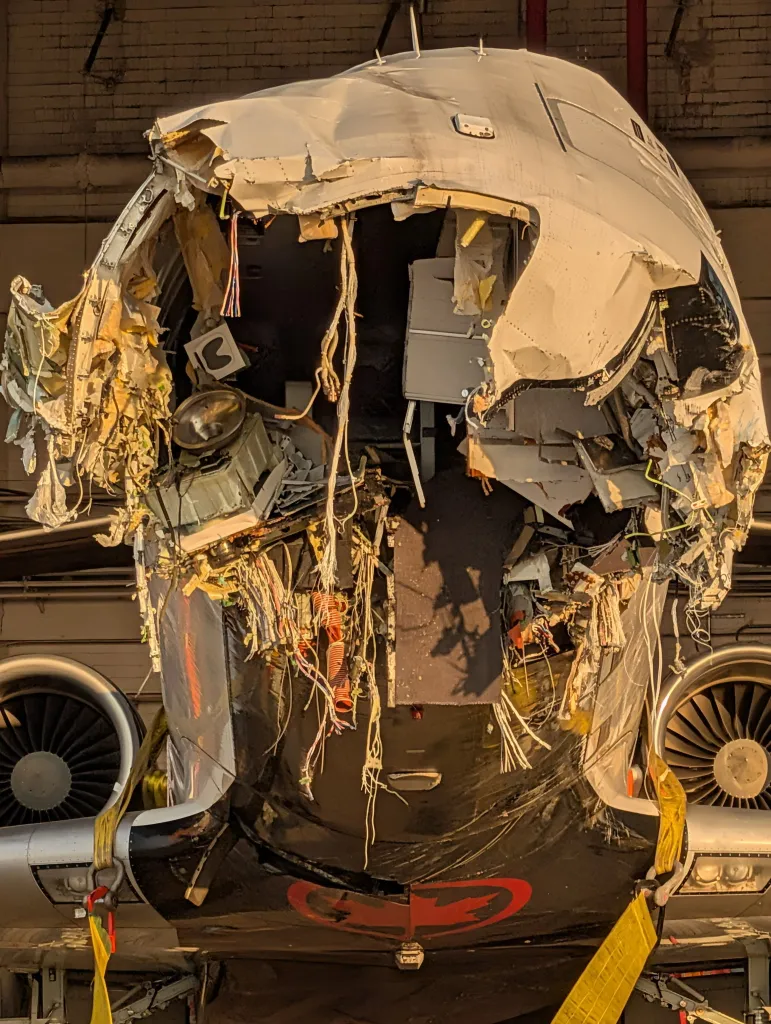 The front fuselage of a damaged Air Canada plane with its nose torn off and internal wiring exposed.
