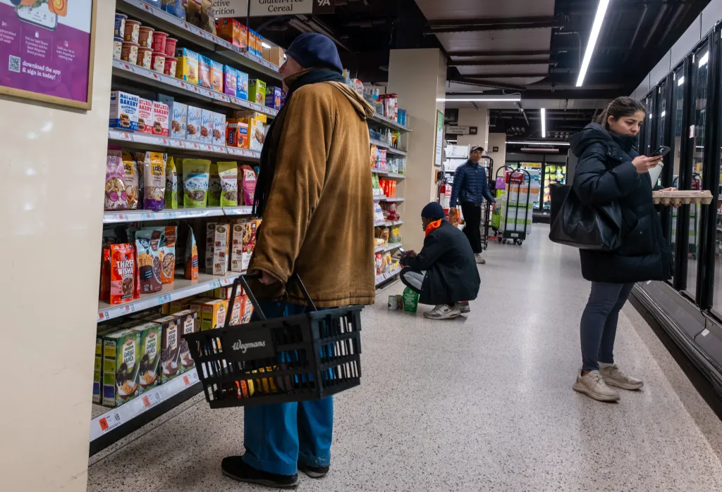 People shopping at a grocery store, with a man in a brown coat holding a black shopping basket in the foreground and shelves of various food products behind him.