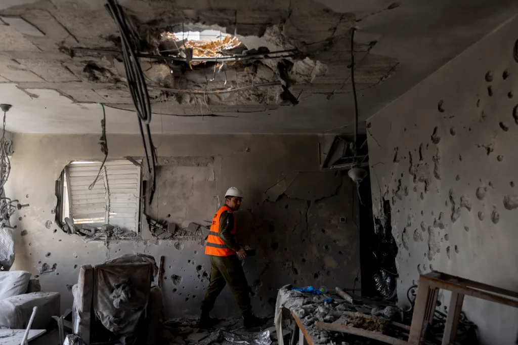 An emergency responder in a hard hat and orange vest inspects the interior of an apartment severely damaged by a missile strike, with debris, holes in the walls, and a large hole in the ceiling.