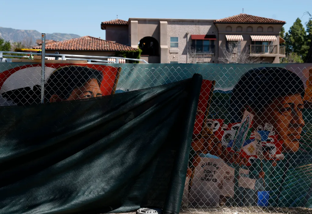 A mural of Cesar Chavez is partially covered by a dark mesh screen on a chain-link fence, with buildings and mountains in the background.