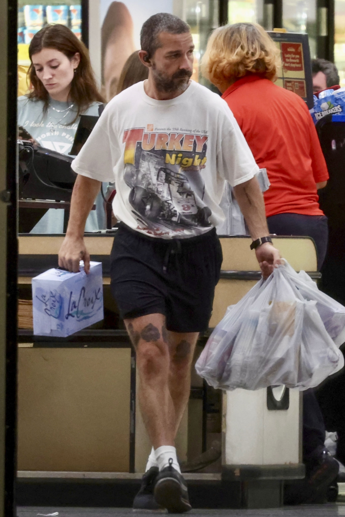 Shia LaBeouf leaves a grocery store carrying La Croix mineral water and plastic bags of groceries.