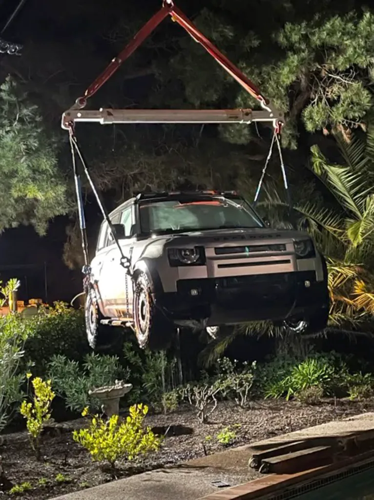 A crane lifts a dark-colored Land Rover Defender SUV from a backyard swimming pool at night.