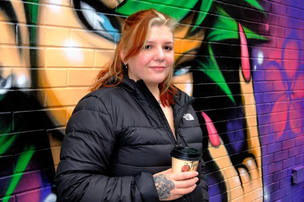 Samantha Vargas, a piercing artist and whistleblower, holding a coffee cup in front of a graffiti-covered brick wall.