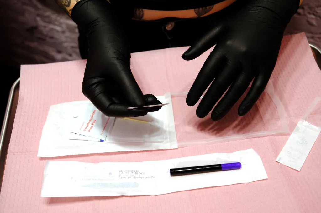 Hands in black gloves prepare piercing tools including a needle, prep pad, and marker on a pink surface.